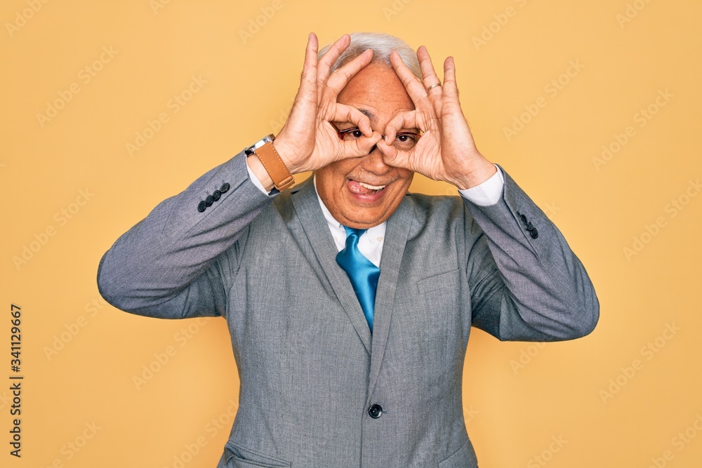 Middle age senior grey-haired handsome business man wearing glasses over yellow background doing ok gesture like binoculars sticking tongue out, eyes looking through fingers. Crazy expression. Middle age senior grey-haired handsome business man wearing glasses over yellow background doing ok gesture like binoculars sticking tongue out, eyes looking through fingers. Crazy expression.