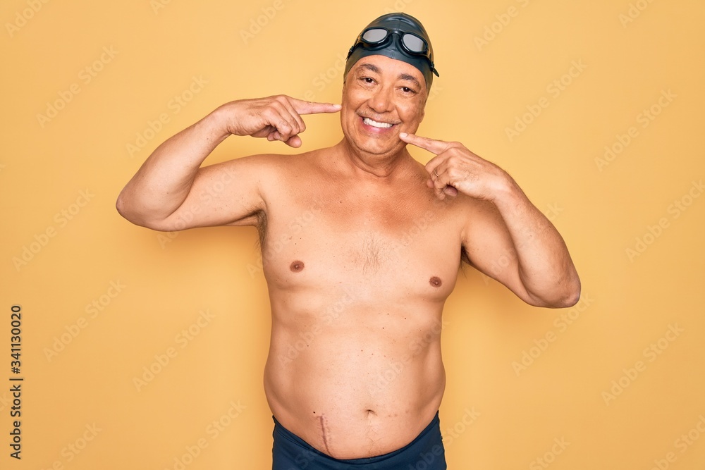 Middle age senior grey-haired swimmer man wearing swimsuit, cap and goggles smiling cheerful showing and pointing with fingers teeth and mouth. Dental health concept.