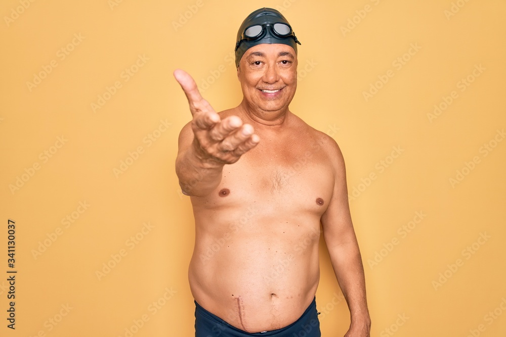 Middle age senior grey-haired swimmer man wearing swimsuit, cap and goggles smiling cheerful offering palm hand giving assistance and acceptance.