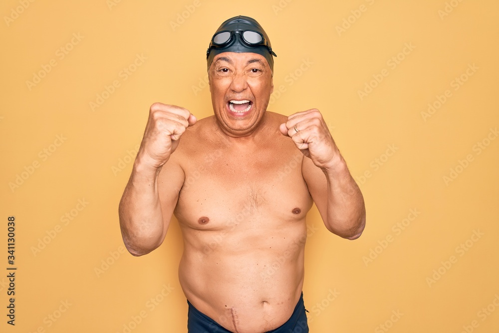 Middle age senior grey-haired swimmer man wearing swimsuit, cap and goggles angry and mad raising fists frustrated and furious while shouting with anger. Rage and aggressive concept.