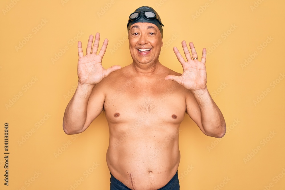 Middle age senior grey-haired swimmer man wearing swimsuit, cap and goggles showing and pointing up with fingers number ten while smiling confident and happy.