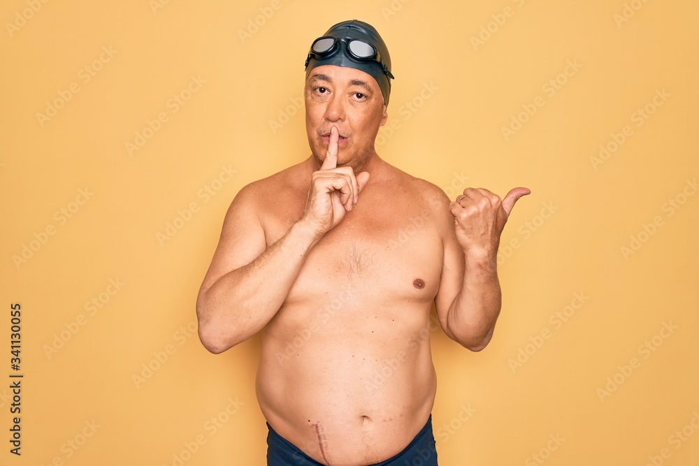 Middle age senior grey-haired swimmer man wearing swimsuit, cap and goggles asking to be quiet with finger on lips pointing with hand to the side. Silence and secret concept.