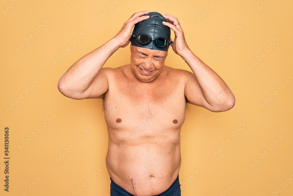 Middle age senior grey-haired swimmer man wearing swimsuit, cap and goggles suffering from headache desperate and stressed because pain and migraine. Hands on head.