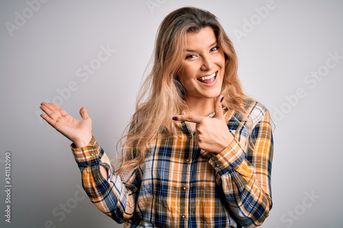 Young beautiful blonde woman wearing casual shirt standing over isolated white background amazed and smiling to the camera while presenting with hand and pointing with finger.