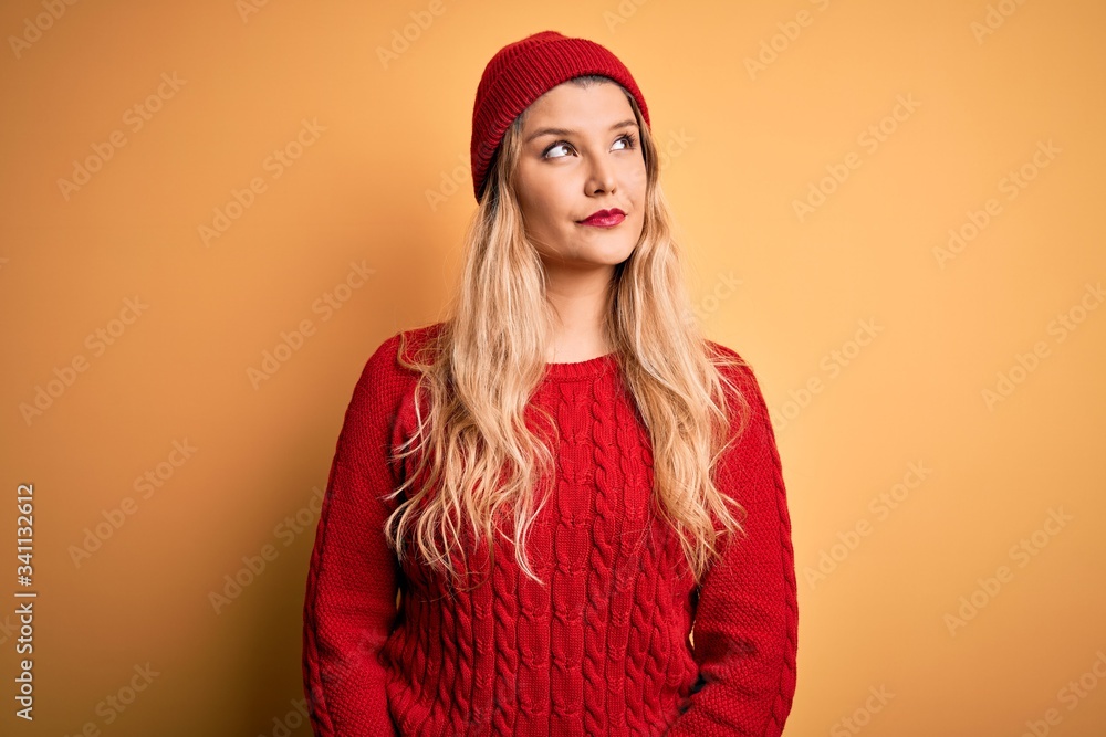 Young beautiful blonde woman wearing casual sweater and wool cap over white background smiling looking to the side and staring away thinking. Young beautiful blonde woman wearing casual sweater and wool cap over white background smiling looking to the side and staring away thinking.