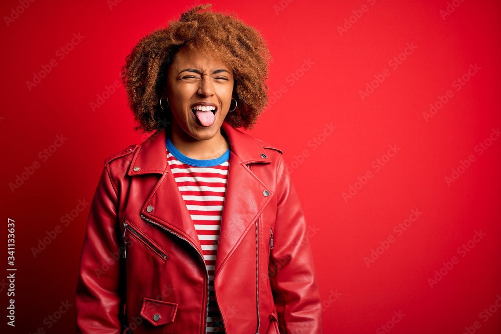 Young beautiful African American afro woman with curly hair wearing casual red jacket sticking tongue out happy with funny expression. Emotion concept.