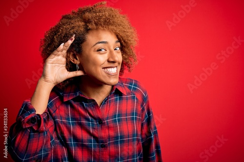 Young beautiful African American afro woman with curly hair wearing casual shirt smiling with hand over ear listening an hearing to rumor or gossip. Deafness concept.