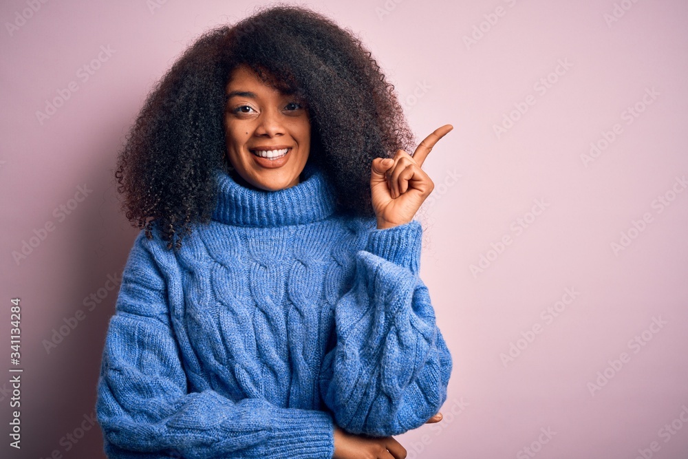 Young beautiful african american woman with afro hair wearing winter sweater over pink background with a big smile on face, pointing with hand and finger to the side looking at the camera. Young beautiful african american woman with afro hair wearing winter sweater over pink background with a big smile on face, pointing with hand and finger to the side looking at the camera.