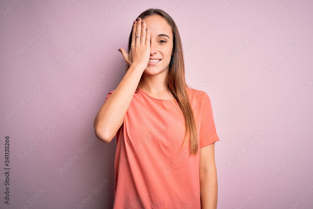 Young beautiful woman wearing casual t-shirt standing over isolated pink background covering one eye with hand, confident smile on face and surprise emotion. Young beautiful woman wearing casual t-shirt standing over isolated pink background covering one eye with hand, confident smile on face and surprise emotion.
