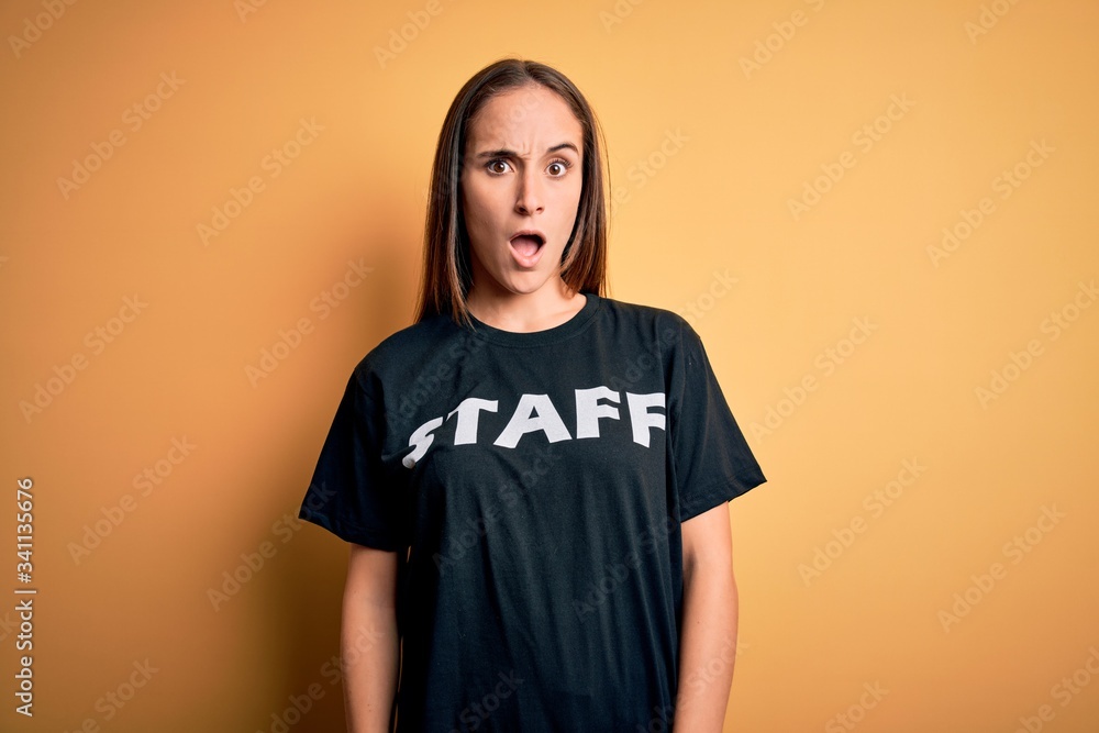 Young beautiful worker woman wearing staff uniform t-shirt over isolated yellow background In shock face, looking skeptical and sarcastic, surprised with open mouth Young beautiful worker woman wearing staff uniform t-shirt over isolated yellow background In shock face, looking skeptical and sarcastic, surprised with open mouth