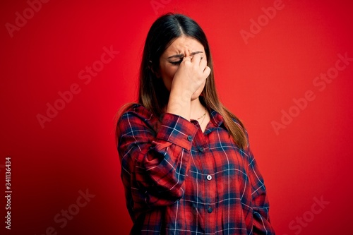 Young beautiful woman wearing casual shirt over red background tired rubbing nose and eyes feeling fatigue and headache. Stress and frustration concept. © Krakenimages.com