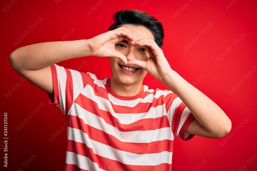 Naklejka premium Young handsome chinese man wearing casual striped t-shirt standing over red background Doing heart shape with hand and fingers smiling looking through sign