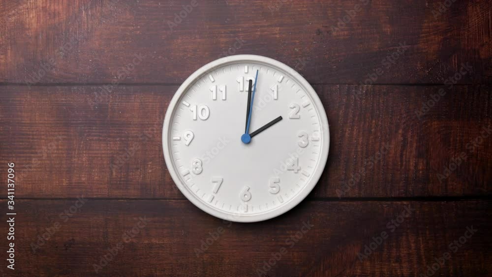 The Time On The Clock Two. White Clock With The Blue Arrow With The Arabic Numerals On An Brown Wooden Background.