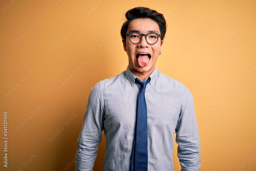 Young handsome chinese businessman wearing glasses and tie over yellow background sticking tongue out happy with funny expression. Emotion concept. Young handsome chinese businessman wearing glasses and tie over yellow background sticking tongue out happy with funny expression. Emotion concept.