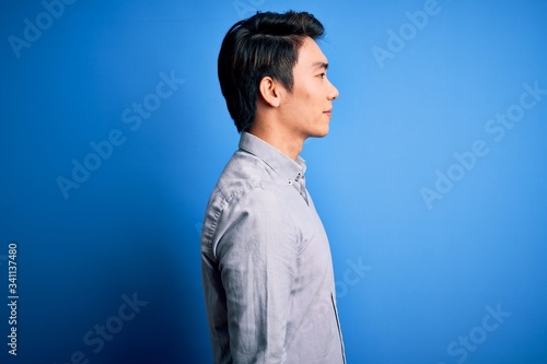 Quadro su tela Young handsome chinese man wearing casual shirt standing over isolated blue background looking to side, relax profile pose with natural face and confident smile