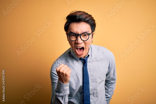 Young handsome chinese businessman wearing glasses and tie over yellow background angry and mad raising fist frustrated and furious while shouting with anger. Rage and aggressive concept.
