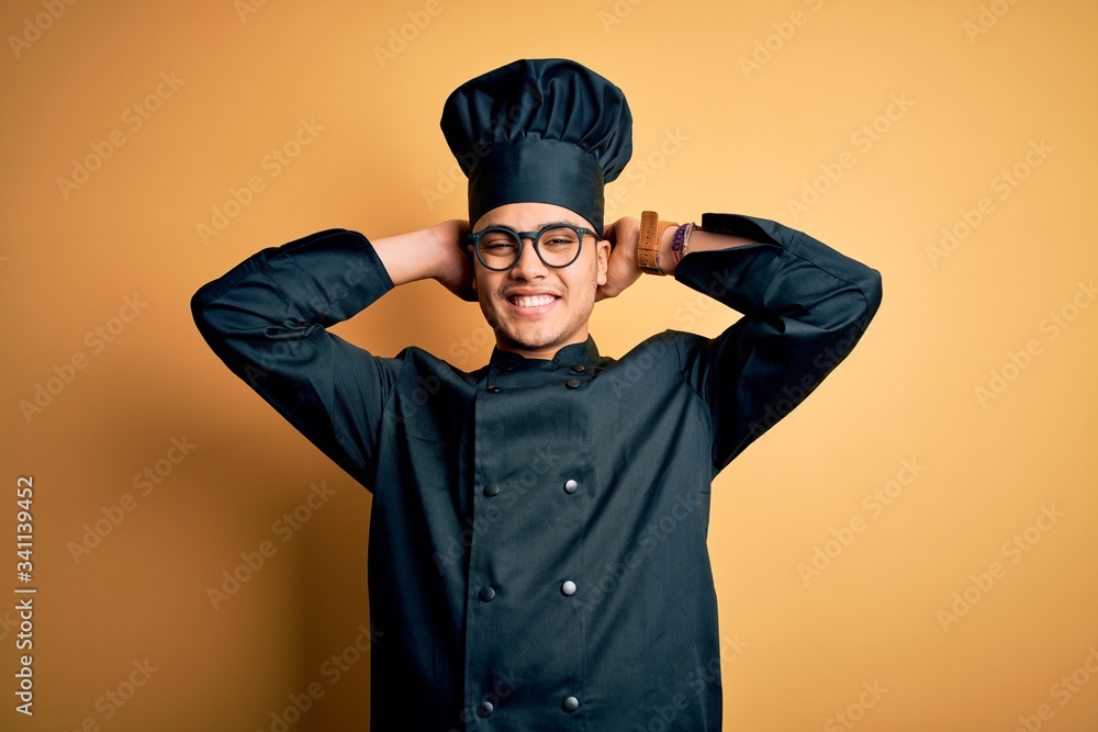 Young brazilian chef man wearing cooker uniform and hat over isolated yellow background relaxing and stretching, arms and hands behind head and neck smiling happy Young brazilian chef man wearing cooker uniform and hat over isolated yellow background relaxing and stretching, arms and hands behind head and neck smiling happy