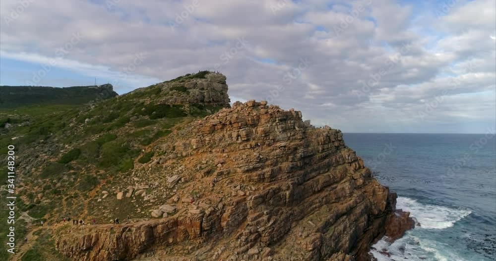 Aerial shot of a parking lot on the shore, people walking to a mountain, drone flying forward while ascending passing over the mountain peak showing a scenic cove - Cape Town, South Africa Aerial shot of a parking lot on the shore, people walking to a mountain, drone flying forward while ascending passing over the mountain peak showing a scenic cove - Cape Town, South Africa