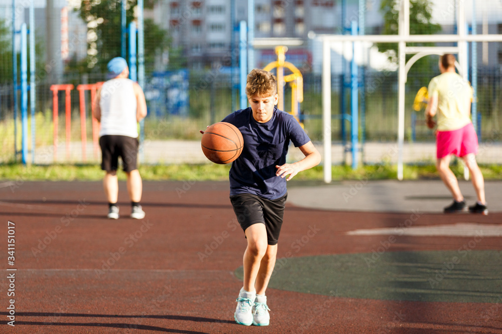 Cute smiling boy in blue t shirt plays basketball on city playground. Active teen enjoying outdoor game with orange ball. Hobby, active lifestyle, sport for kids.