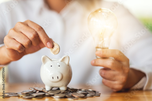 Slika na platnu A woman holding light bulb while putting coins into piggy bank for saving money