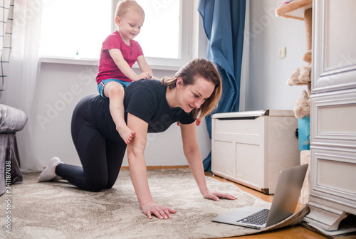Beautiful young woman in a black tracksuit doing workout exercises at home watching videos on her laptop but the child prevents her from doing