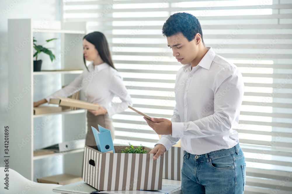 Upset young brunette male packing his personal stuff into box, his female colleague standing near closet Upset young brunette male packing his personal stuff into box, his female colleague standing near closet