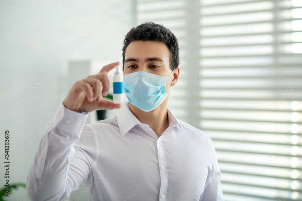 Dark-haired man in a protective mask holding a disinfector in his hand an looking serious