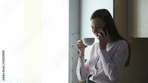 A young girl standing by a window at Khuong and talking on the phone and drinking a cup of coffee. Business woman is going to work. photo