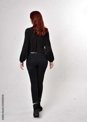 Portrait of a pretty girl with red hair wearing black jeans, boots and a blouse. full length standing pose on a studio background, with back to the camera. 