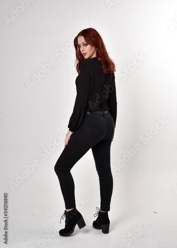 Portrait of a pretty girl with red hair wearing black jeans, boots and a blouse. full length standing pose on a studio background, with back to the camera. 