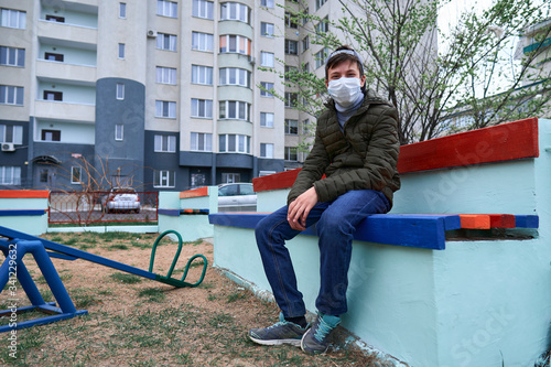 Fototapeta Naklejka Na Ścianę i Meble -  teen boy sits on a bench on playground near high-rise buildings with apartments, a medical mask on his face protects against viruses and dust