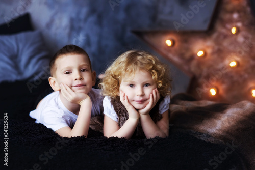 Two children, boy and girl, brother and sister, siblings, laying in bed in loft styled bedroom with star on background. Curly blonde girl and brother. Kids spend time together. Family. Self Isolation