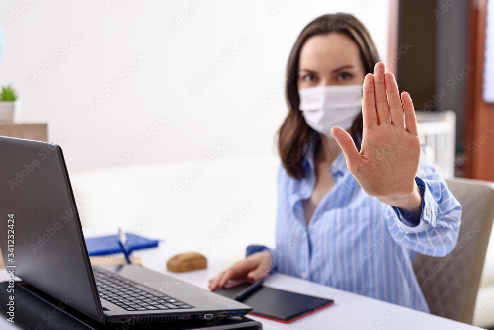 woman with medical mask on face shows stop with hand while sitting at laptop, remote work in quarantine, keeping distance concept woman with medical mask on face shows stop with hand while sitting at laptop, remote work in quarantine, keeping distance concept
