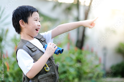 Curious little young Asian boy holding a binoculars and point finger out in the garden. Exploring the world, outdoors activity. Feeling fun. Boy wear green outdoors vest. Holiday, education concept