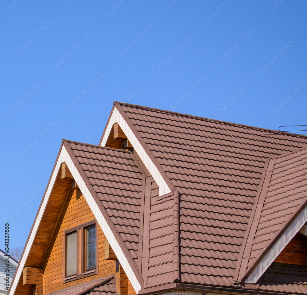 Gable triangular roof with a large pediment. Glued beams with grooves, attic window, brown tiles. Blue sky background. Gable triangular roof with a large pediment. Glued beams with grooves, attic window, brown tiles. Blue sky background.