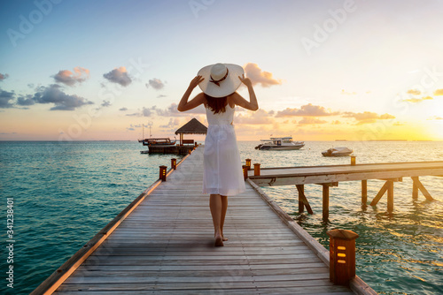 A beautiful tourist woman in white dress on summer vacations walks over a wooden pier into the tropical sunset on the Maldives islands