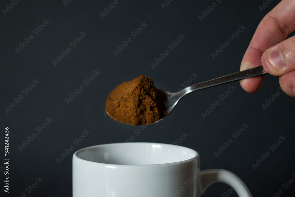 Close up of hand pouring coffee from spoon to white coffee mug, cup on black background Close up of hand pouring coffee from spoon to white coffee mug, cup on black background
