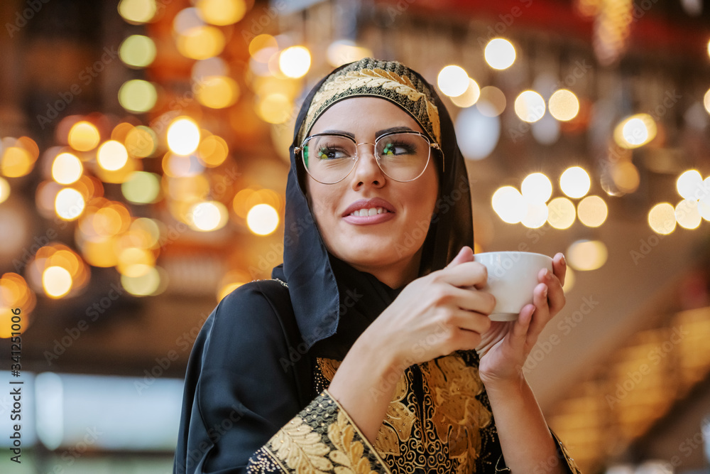 Φωτογραφία Attractive positive smiling muslim woman in traditional wear sitting in cafe alone, holding cup of fresh coffee and looking away
