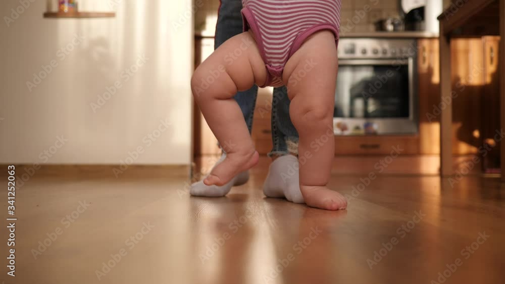 Mother supports toddler baby child makes first steps barefoot on the floor at home Mother supports toddler baby child makes first steps barefoot on the floor at home