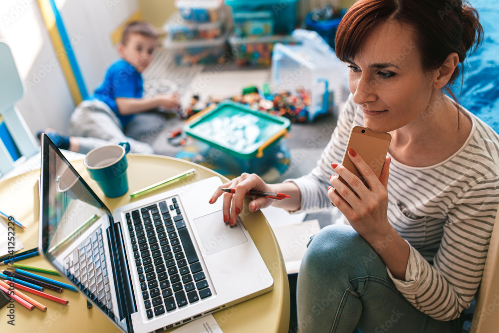 Mother working remotely on laptop while taking care of her son playing with toys in his room. Mother working remotely on laptop while taking care of her son playing with toys in his room.