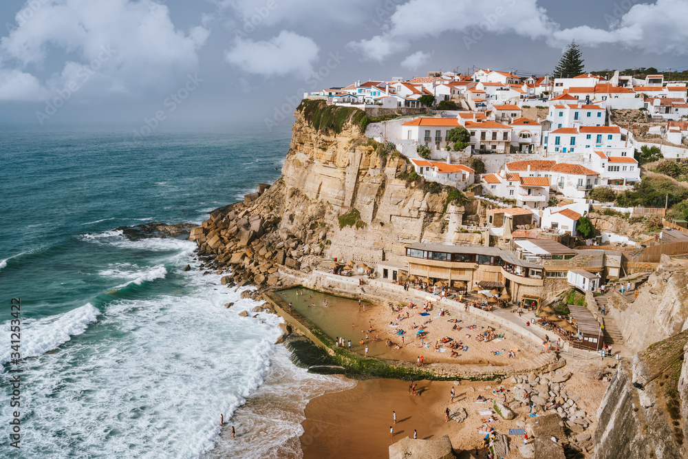 Holiday vibes on beautiful Azenhas do Mar village and wave protected beach. White chalk houses on the edge of a cliff. Bly sky and white clouds. Sintra Landmark, Portugal, Europe