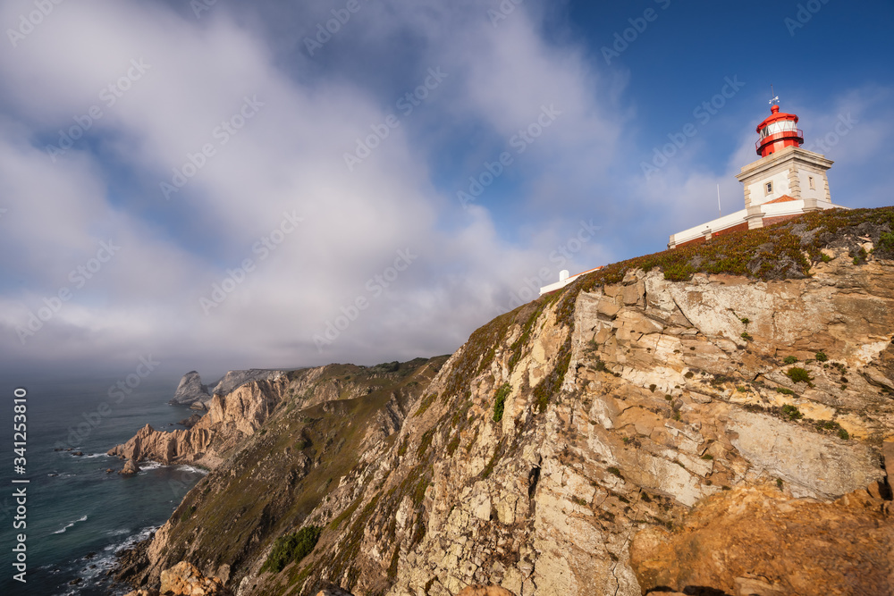 Cabo da Roca Lighthouse at cliff edge in evening sunlight against beautiful cloudscape. Most westerly point of the Europe mainland, Sintra, Portugal. Holiday travel destination