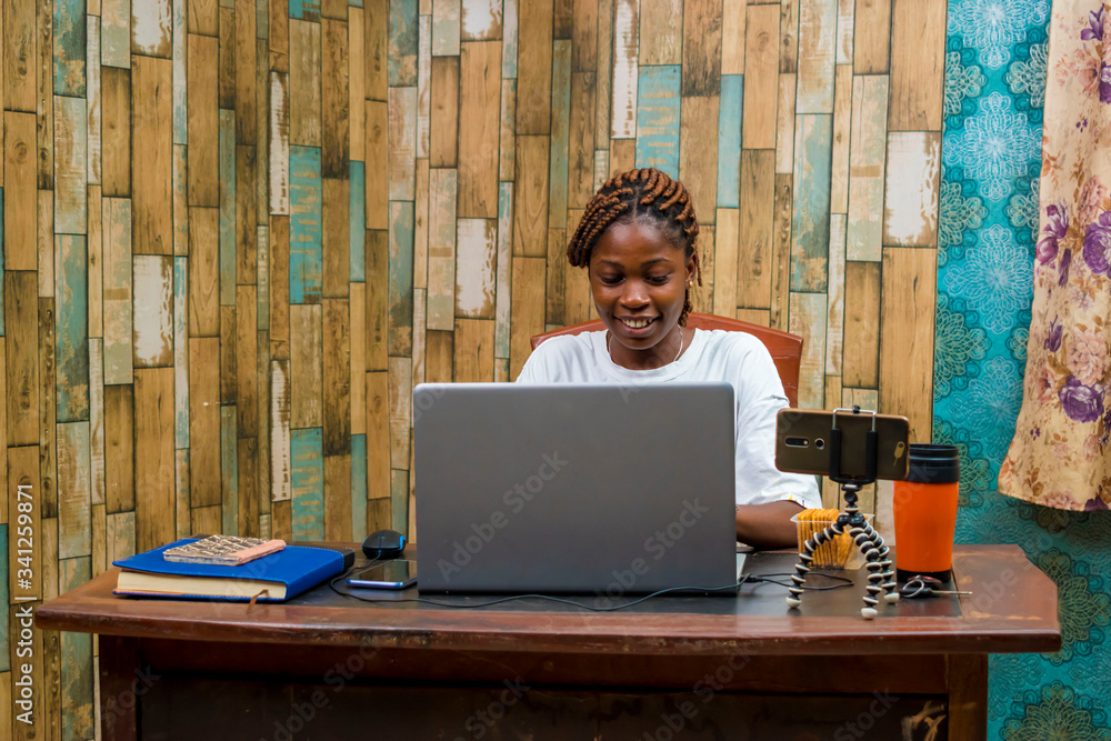 young black woman sitting at home, vlogging with her smartphone while working on her laptop young black woman sitting at home, vlogging with her smartphone while working on her laptop