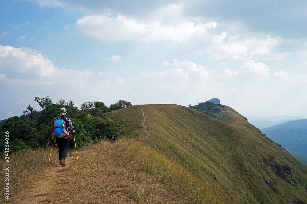 A girl trekking along grassy pathway among green mountain hill and cloudy blue sky