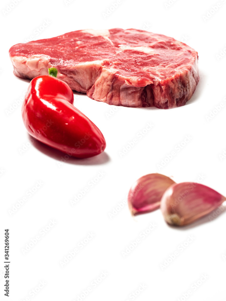 Fresh raw rib eye steak in focus, red chilli pepper and garlic cloves out of focus. White isolated background. Fresh raw rib eye steak in focus, red chilli pepper and garlic cloves out of focus. White isolated background.