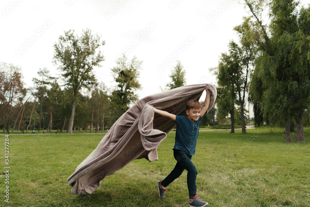 Preschool boy running with a flying blanket on green grass in the park on a summer day Preschool boy running with a flying blanket on green grass in the park on a summer day