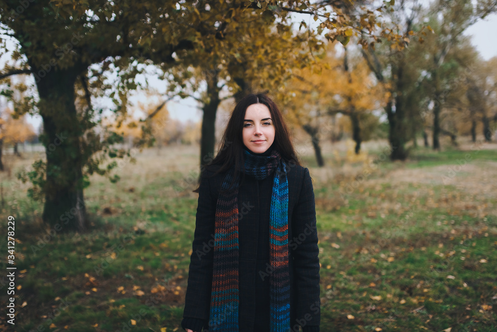 A young girl with dark hair in an autumn coat and a knitted scarf stands in a park among autumn trees. Walking in the fresh air ao quarantine. A young girl with dark hair in an autumn coat and a knitted scarf stands in a park among autumn trees. Walking in the fresh air ao quarantine.