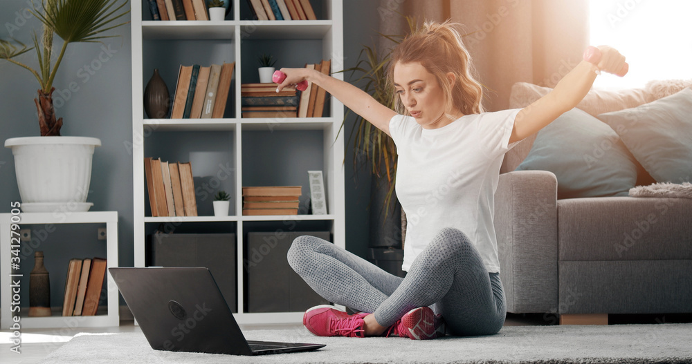 Young lady sitting on floor watching online video on laptop and repeating fitness exercises at home