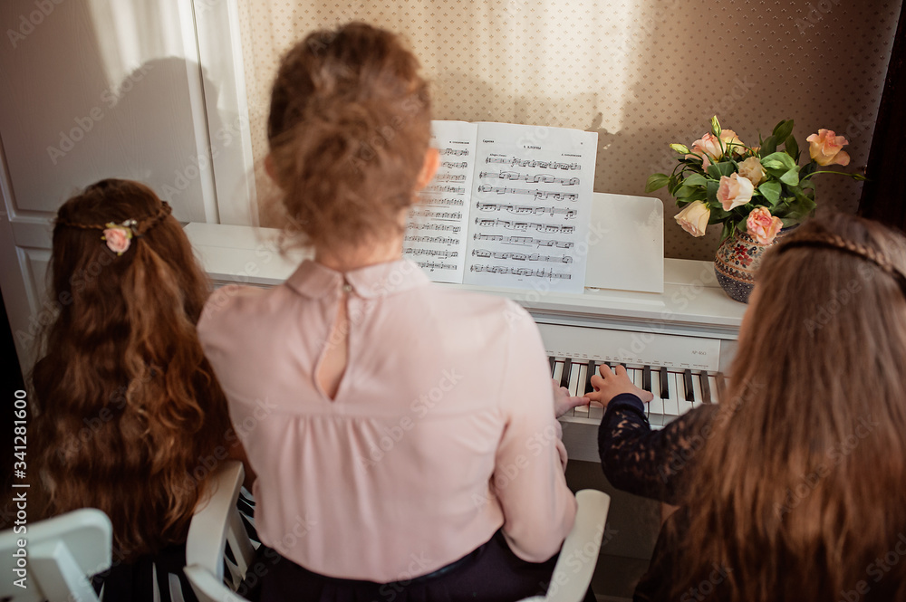 Home piano lesson. A woman and two girls practice sheet music on one musical instrument. Family concept. The idea of activities for children during quarantine. Home piano lesson. A woman and two girls practice sheet music on one musical instrument. Family concept. The idea of activities for children during quarantine.