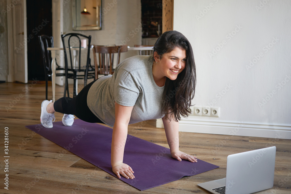 Sporty plus size woman in sportswear working out at home, doing plank on yoga  mat in front of open laptop, repeating instructions by professional fitness  trainer watching online video tutorial Stock Photo